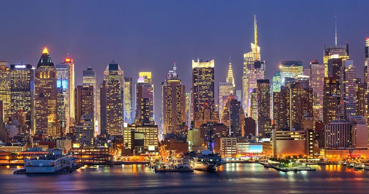 City skyline at dusk with illuminated skyscrapers reflecting on the water.