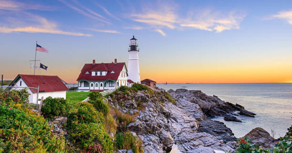 Lighthouse and white buildings on rocky coastline at sunset with calm ocean and pastel sky