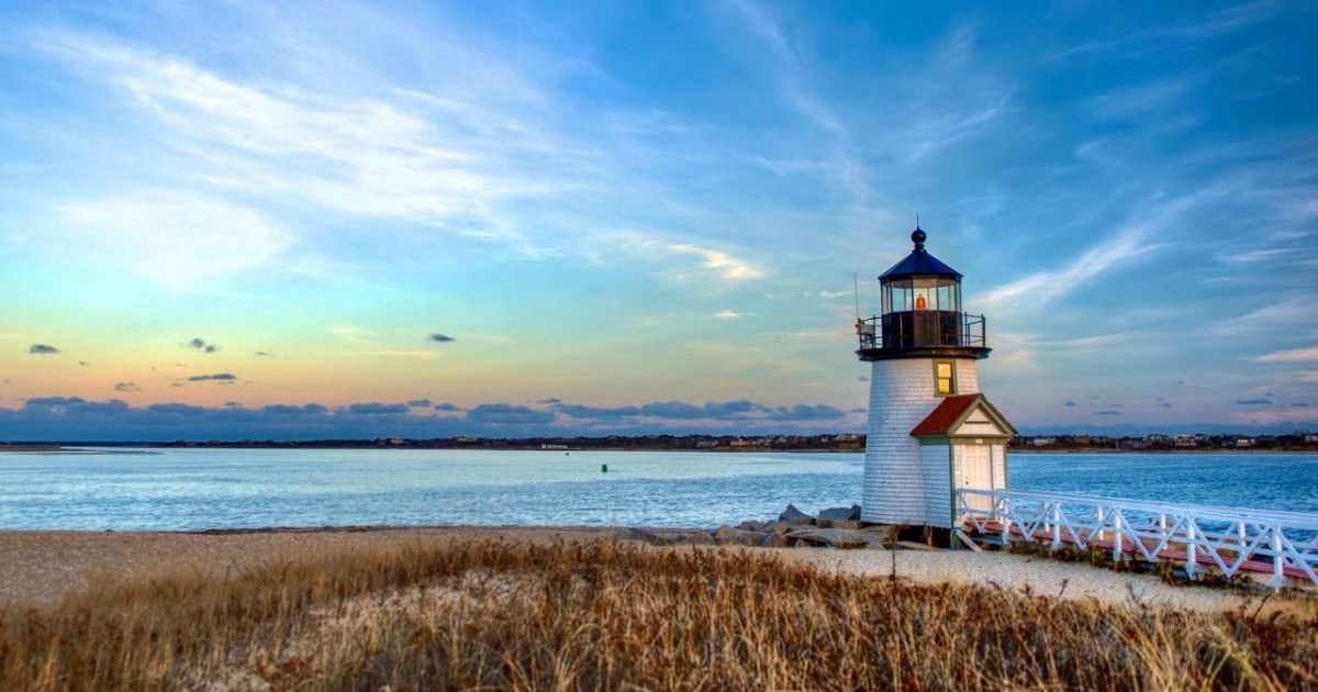Lighthouse on a coastal shoreline at sunset, with calm water, grassy foreground, and dramatic blue sky.