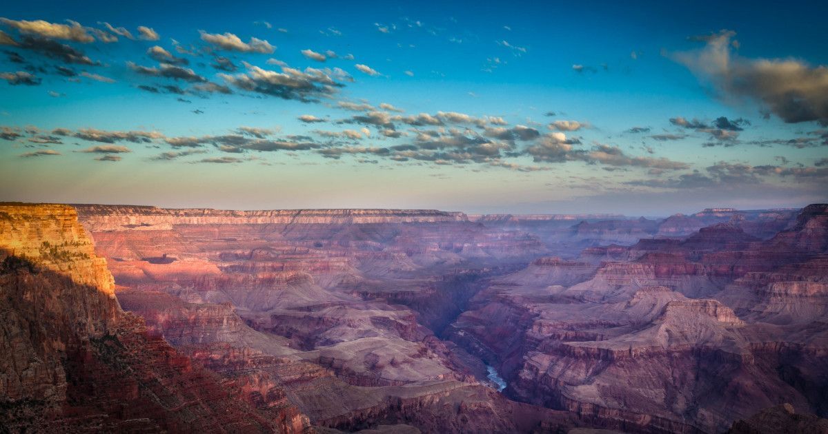 Wide view of the Grand Canyon at sunset with layered cliffs, a blue sky, and pink-orange light