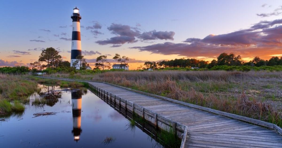 Lighthouse at sunset beside a marsh, with a wooden boardwalk and water reflecting the sky.
