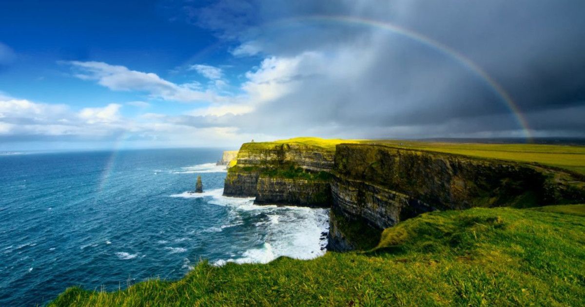 Green cliff overlooking a blue ocean with waves and a rainbow under a cloudy sky