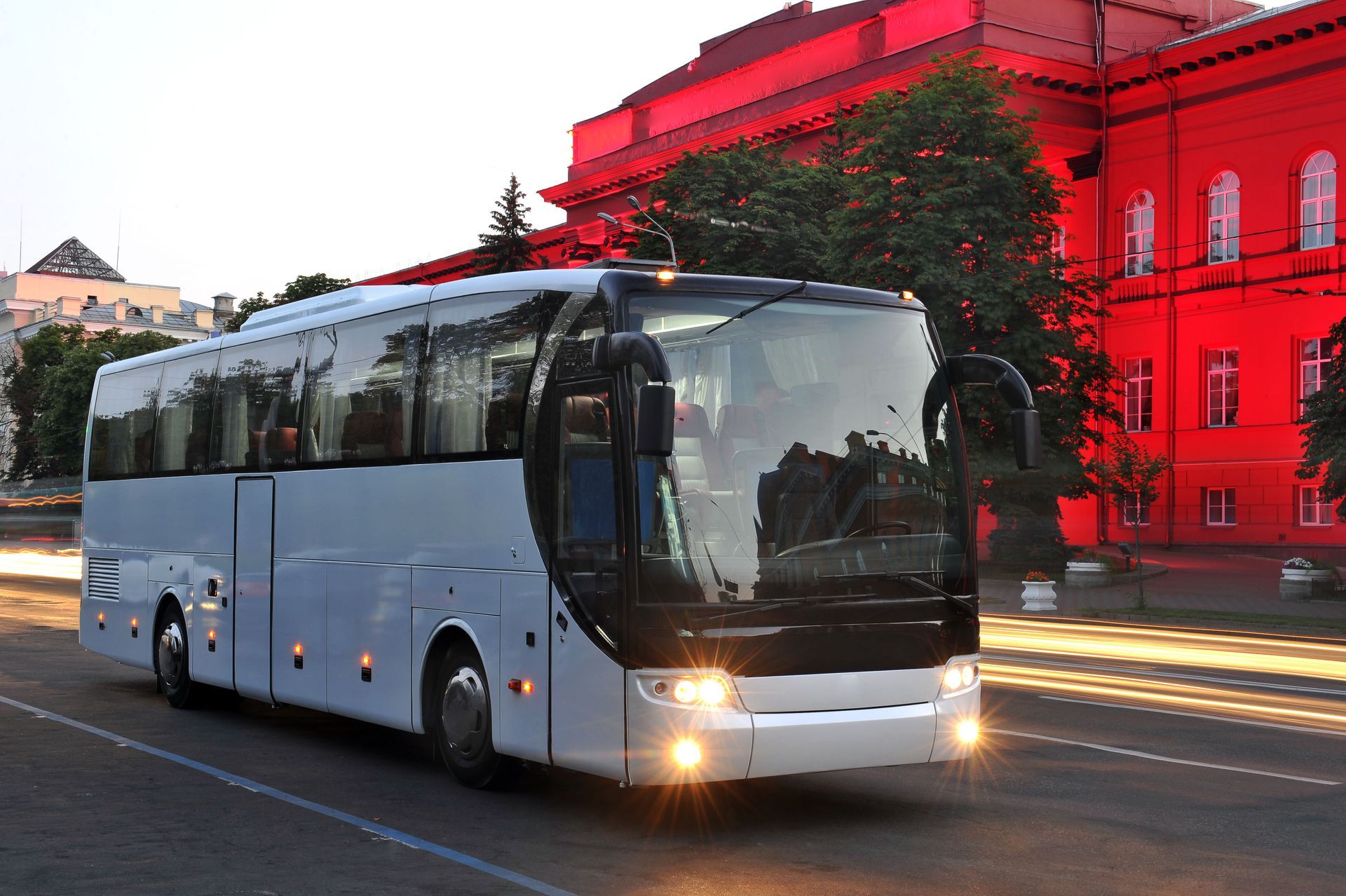 White coach bus driving past a red-lit building at dusk.