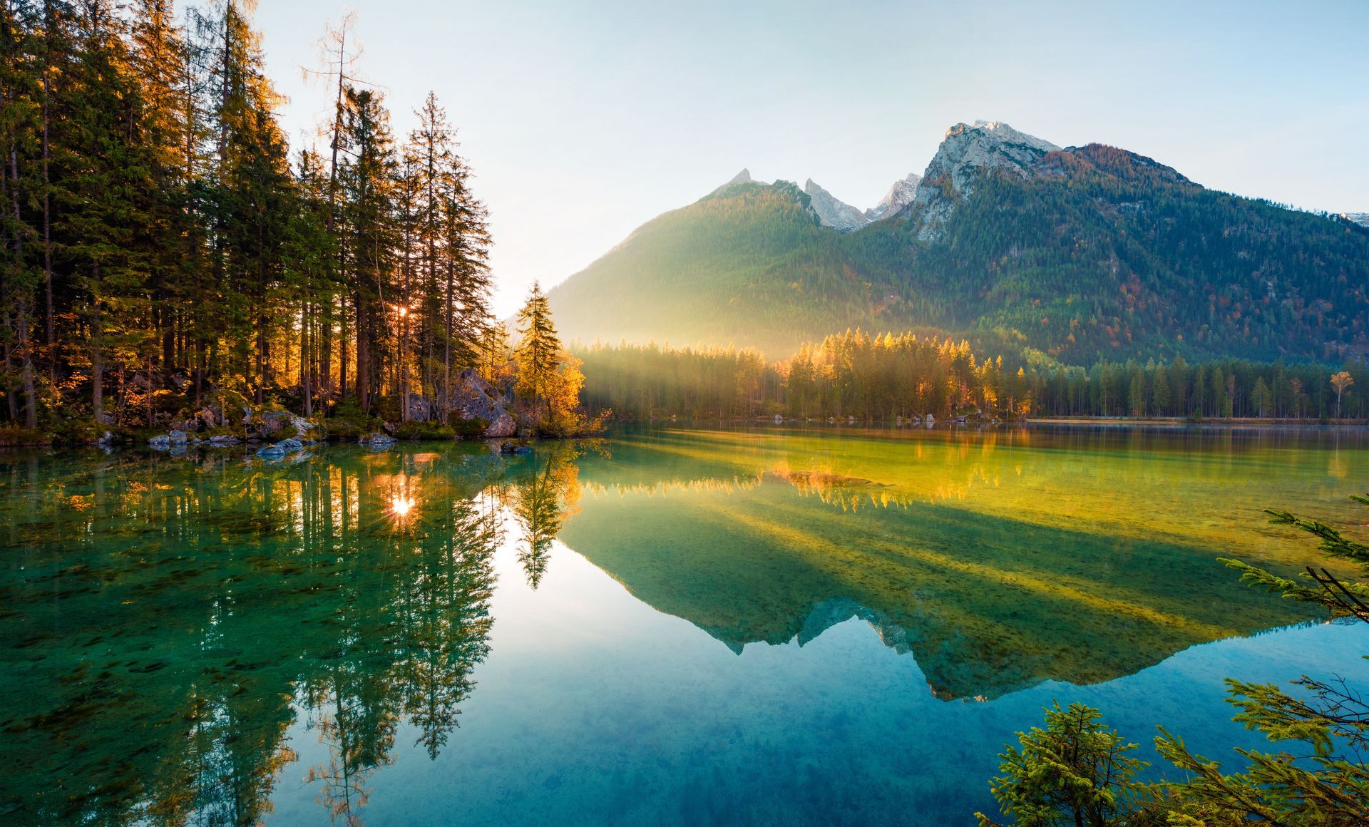 Mountain lake at sunrise with pine trees, green reflections, and snow-dusted peaks