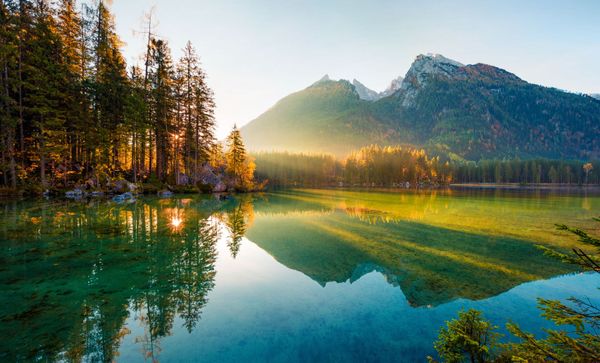 Mountain lake at sunrise with pine trees, green reflections, and snow-dusted peaks