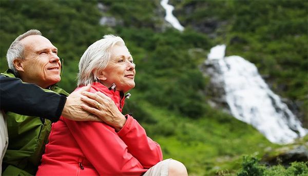 Two hikers in colorful jackets sit by a waterfall in a lush green forest