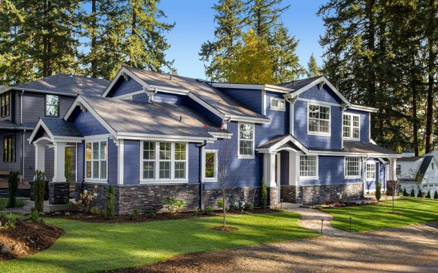 Large blue-gray suburban house with white trim, stone accents, and a manicured lawn under tall trees