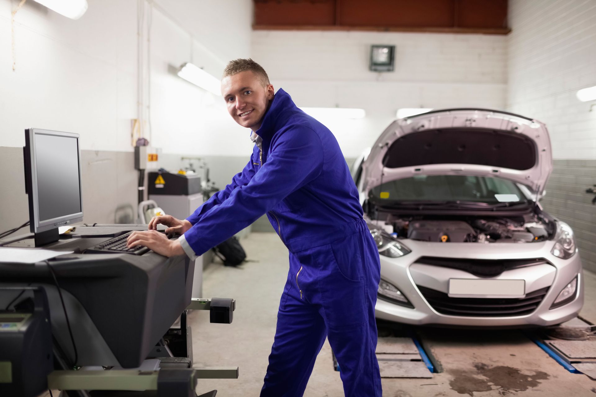 Mechanic in blue coveralls using a computer beside a car with its hood open in a repair shop