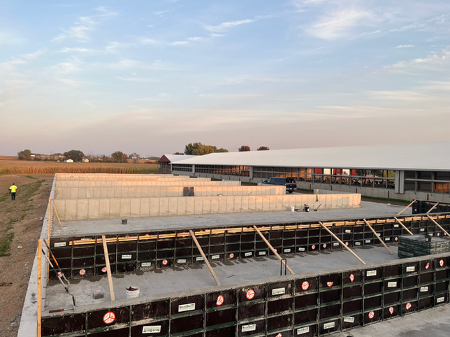 Construction site showing concrete walls under construction and a long barn structure in a rural field at sunset.