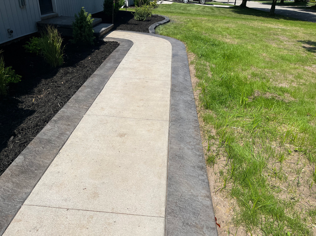 A concrete walkway with dark gray textured borders, winding through a yard with black mulch beds and a lawn.