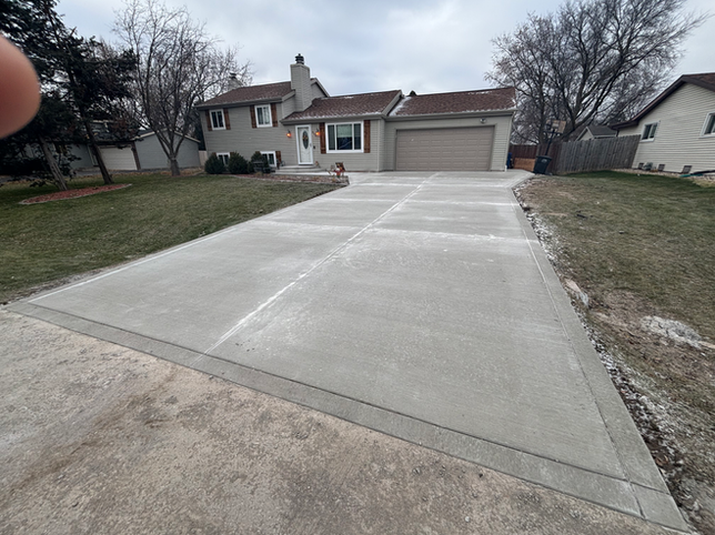 A newly poured concrete driveway leads to a suburban house with a tan garage and a brick-accented front window.