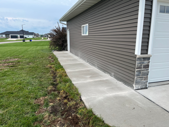 A concrete sidewalk runs along the side of a grey house with brown siding and stone detailing near a garage door.