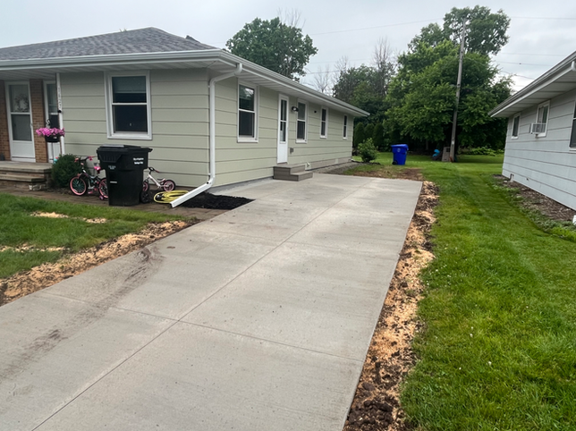 A newly poured concrete driveway leads to the side entrance of a light green house with a white downspout.