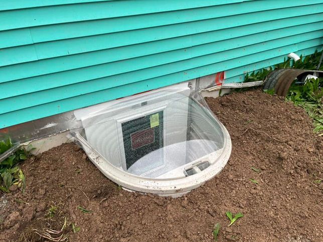 A basement window well with a clear plastic cover set against the turquoise siding of a house, surrounded by loose soil.