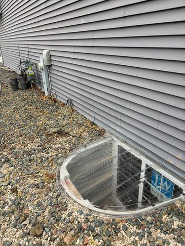 A window well with a metal grate sits next to the gray-sided wall of a house, surrounded by gravel ground.