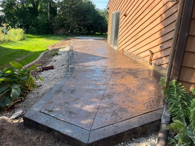 Stamped concrete patio with a dark border, extending alongside a brown-sided house with landscaping and a gravel border.