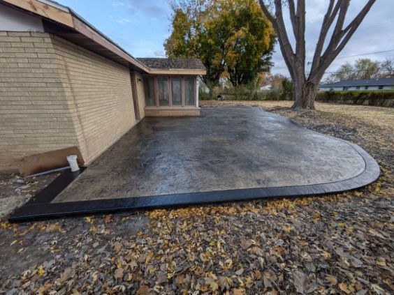 A newly installed, stamped concrete patio with a dark border alongside a tan brick house on an autumn day.
