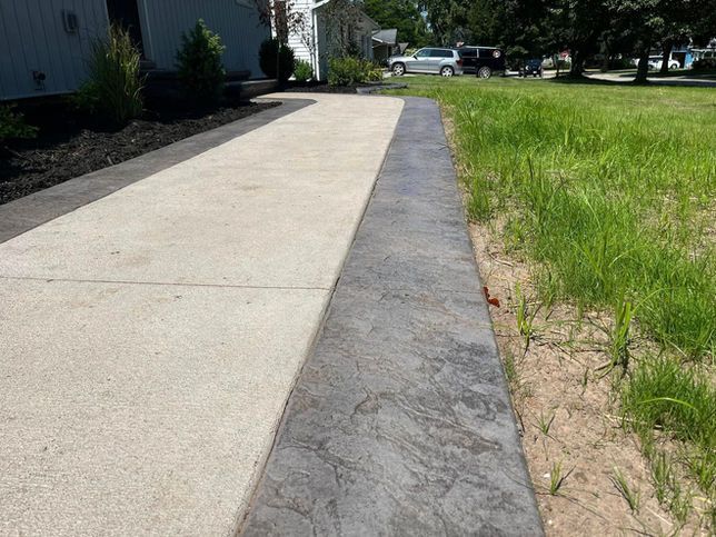 A sidewalk with a smooth concrete center and a textured, dark gray concrete border beside a grassy lawn.