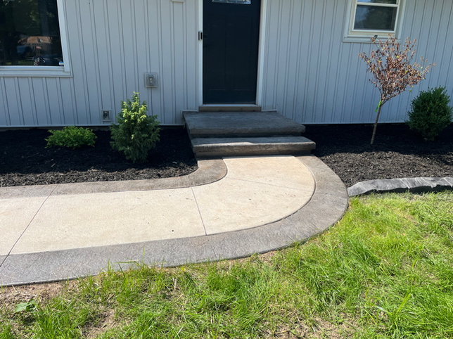 Concrete walkway with a curved border leading to two steps and a black front door of a house with light grey siding.