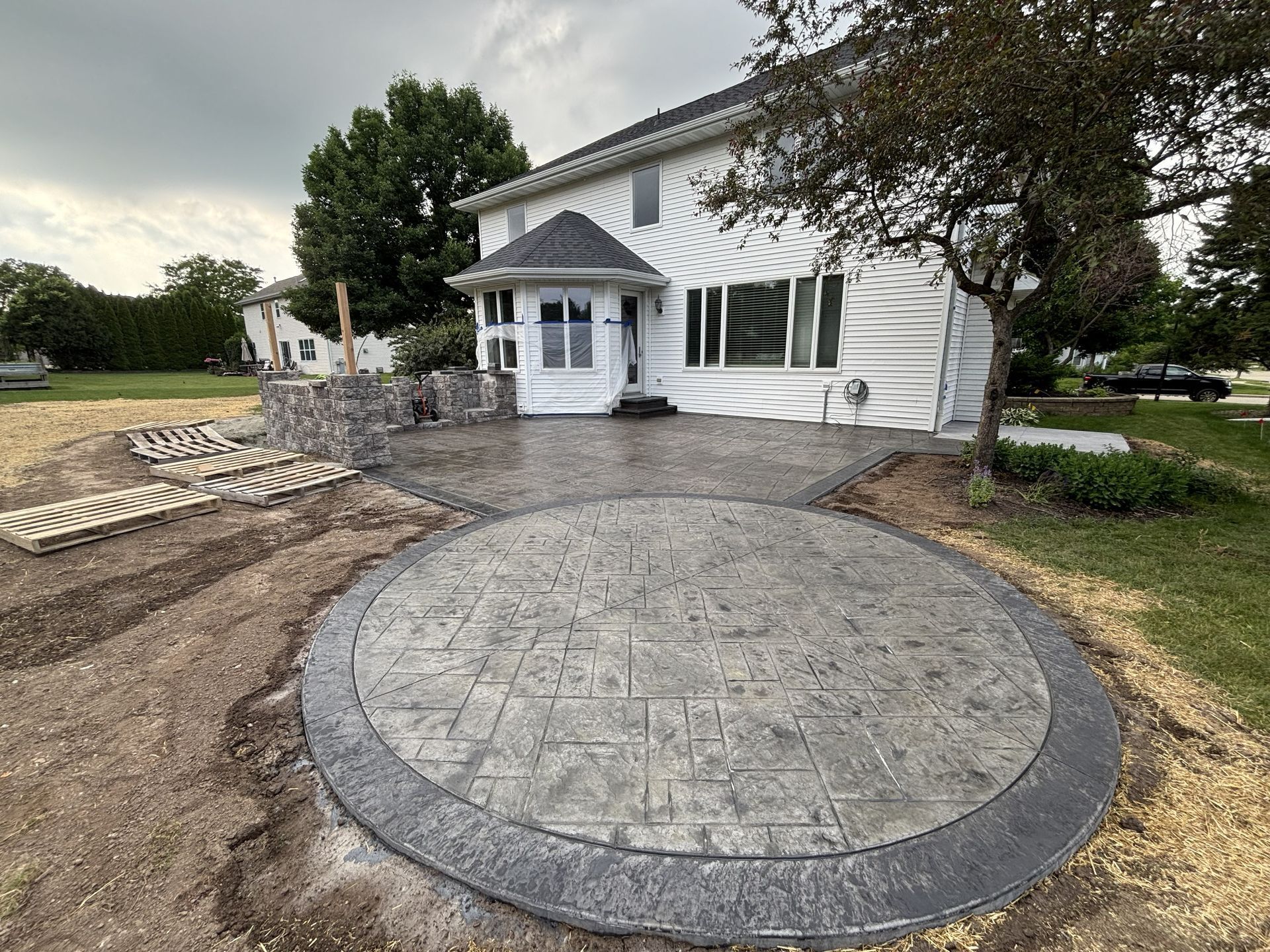 A newly installed, circular stamped concrete patio with a dark border next to a white house with a green lawn.