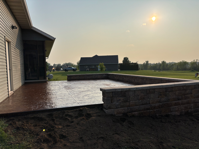A tan house with a screened porch and a large stamped concrete patio featuring a tiered stone retaining wall at sunset.