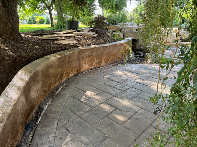 A curved, tan concrete retaining wall lines a patio with patterned grey pavers in a sunny backyard garden setting.