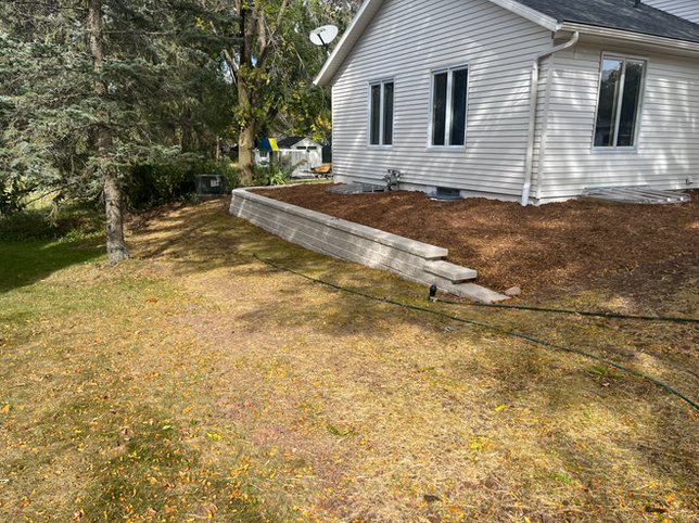 A beige stone retaining wall sits in front of a white house with dark windows, surrounded by wood mulch and grass.