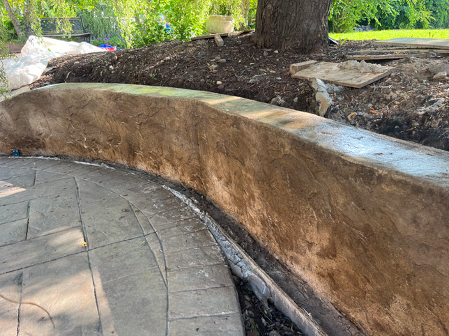 A curved, textured concrete retaining wall stands next to a stamped concrete patio in a yard with a tree in the background.