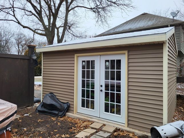 A brown-sided shed with double French doors, standing in a backyard next to a fence on a cloudy day.