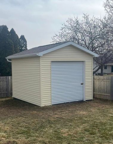 A small, detached cream-colored vinyl shed with a grey metal roll-up door and a shingled roof, set in a fenced yard.