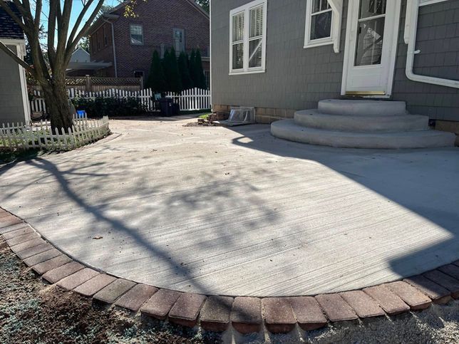 A light gray concrete patio with a curved brick border leading to a set of three semicircular concrete steps at a house.