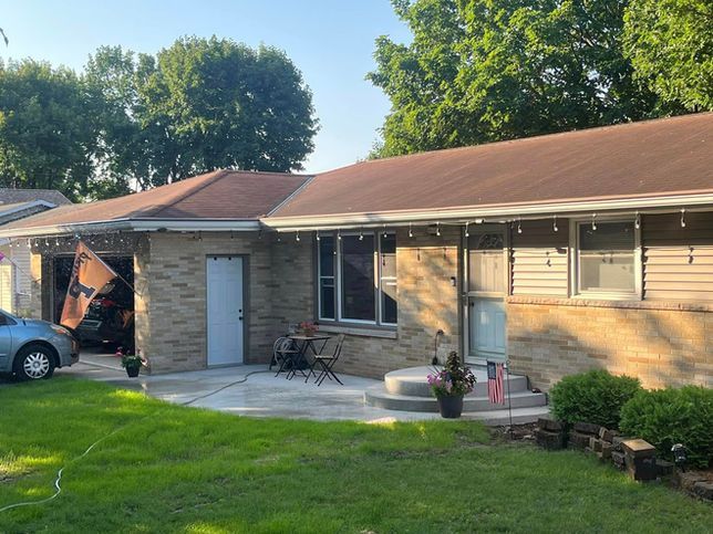 Single-story house with a stone facade, a patio, a detached garage, a parked car, and a green front yard on a sunny day.