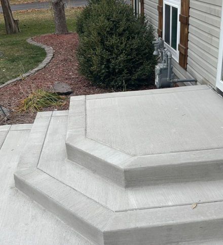 Concrete front steps with a multi-sided design leading to a house with beige siding and a nearby evergreen bush.