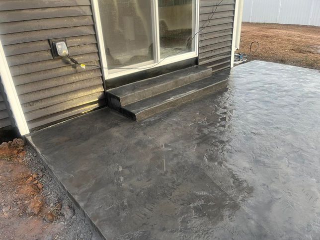 A newly poured dark gray stamped concrete patio with two steps leading up to a sliding glass door on the side of a house.