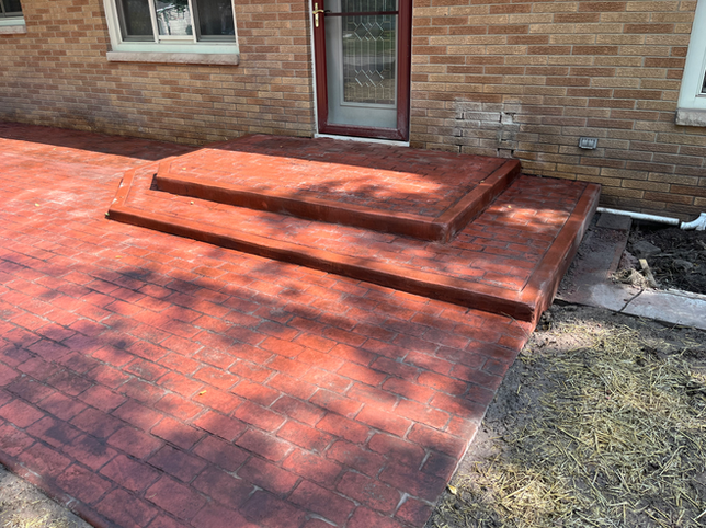 A red, brick-patterned concrete patio and porch with two steps leading to a house entrance.