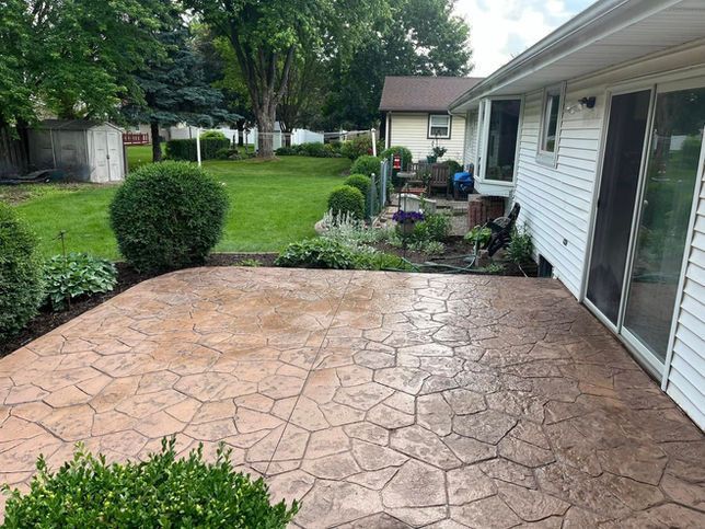 A stamped concrete patio featuring a tan, flagstone pattern in a backyard with a lawn, landscaping, and a house exterior.