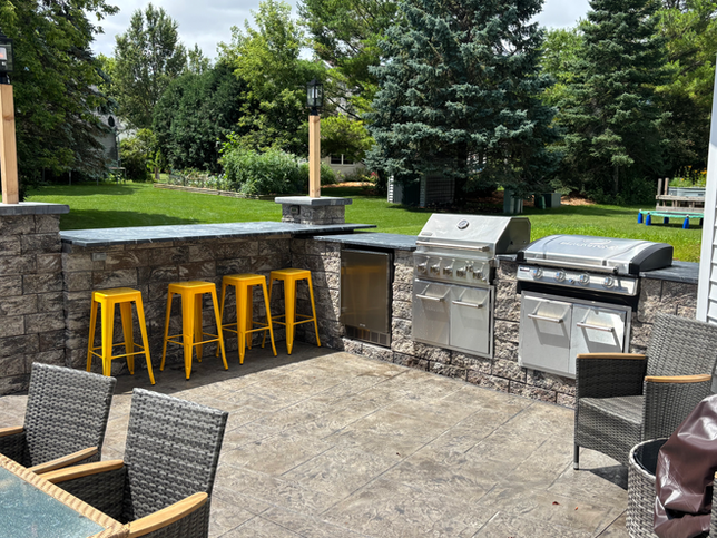 An outdoor kitchen featuring stone masonry, a stainless steel grill, a bar counter with four yellow stools, and patio chairs.
