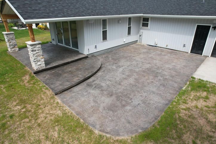 A gray, stamped concrete patio with a curved step, attached to the back of a light gray house with stone support pillars.