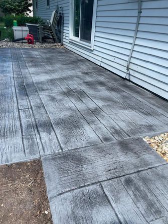 A gray, wood-grain patterned stamped concrete patio outside a house with white siding and a sliding glass door.