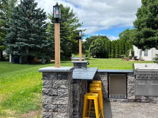 Outdoor kitchen with a stone counter, stainless grill, and yellow stools under two wooden lamp posts on a grassy lawn.