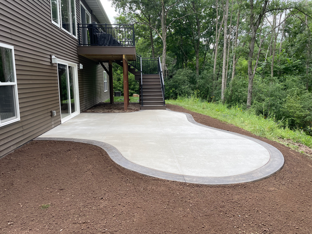 A newly poured concrete patio with a dark paver border, located behind a house next to wooden stairs and a lush tree line.