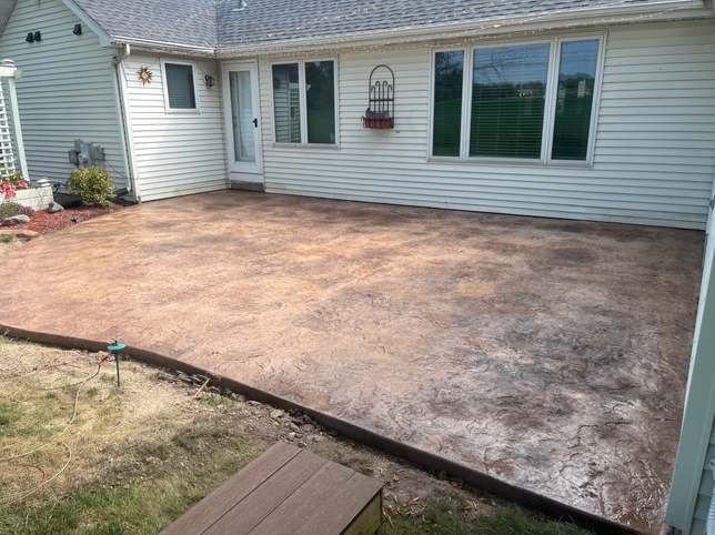 A rectangular stamped concrete patio with mottled brown tones attached to the back of a house with light beige siding.