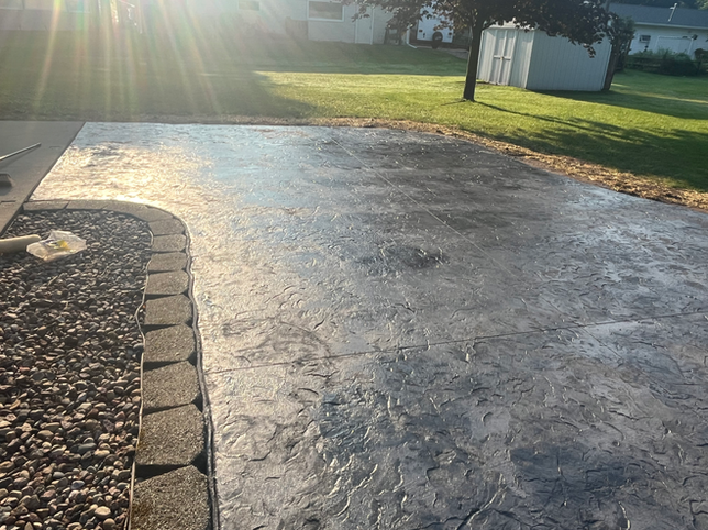 A stamped concrete patio bordered by gray stone pavers and a bed of landscaping rocks in a sunny backyard.