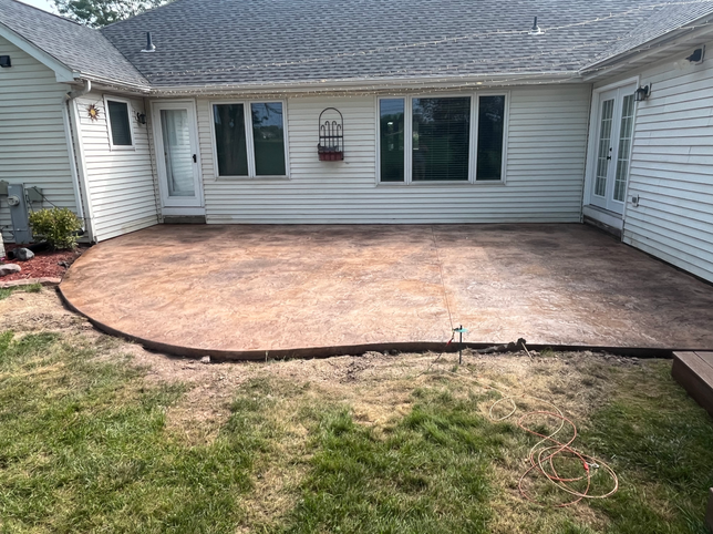 A stamped, textured concrete patio with brown and tan hues sits behind a light-colored house with a grassy lawn.