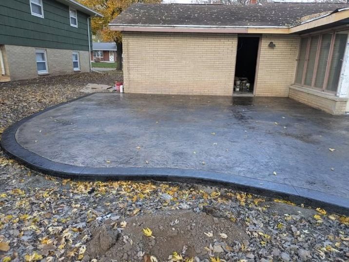 A new curved, dark gray stamped concrete patio sits beside a brick house with a gravel yard in the foreground.
