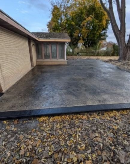A stamped, dark-gray concrete patio adjacent to a tan brick house, with a dark border and scattered fallen leaves nearby.