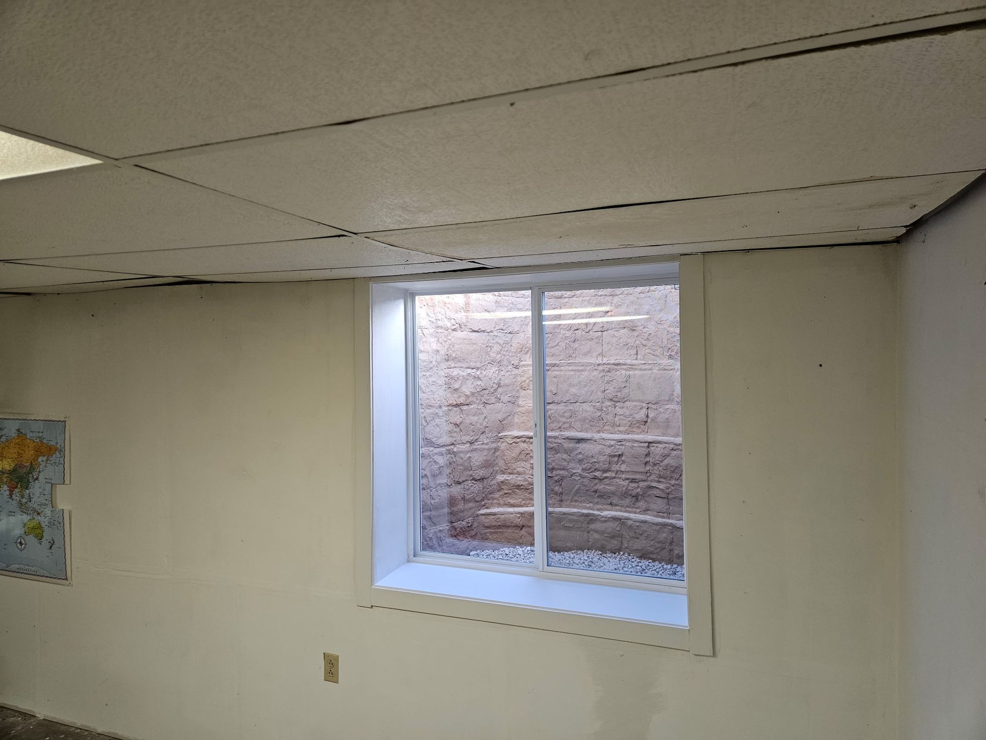 An interior basement wall with a white-framed window looking out into a window well with a stone wall and gravel.