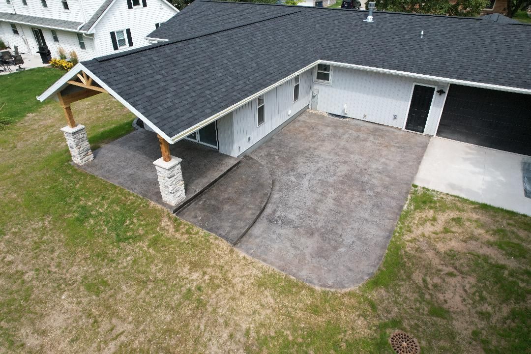A high-angle view of a white home with a black roof, a covered porch with stone pillars, and a gray concrete patio.