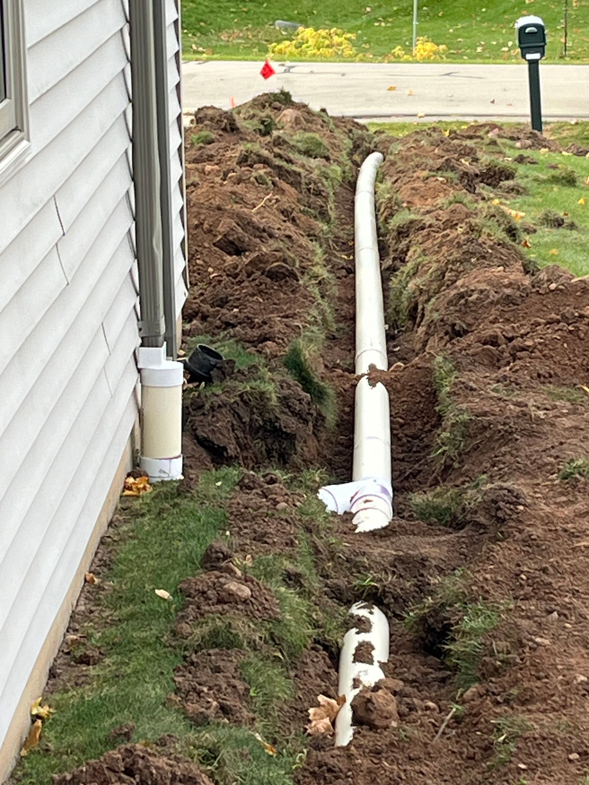 A PVC drainage pipe runs through a freshly dug trench along the side of a house with light-colored siding.