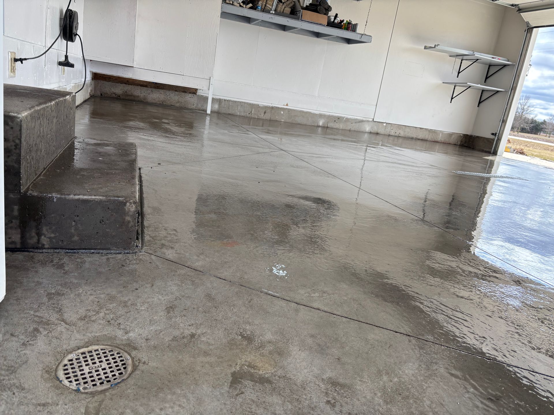 A wet concrete garage floor with a drain in the foreground, steps on the left, and storage shelving on the back walls.
